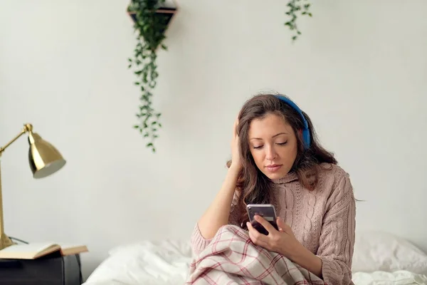 Młoda, wesoła kobieta leżąca na łóżku tasując piosenki przez telefon komórkowy. Zrelaksowana dziewczyna słuchająca muzyki ze słuchawkami w domu. Happy woman in casual korzystających z muzyki w sypialni podczas smsowania wiadomości telefon. — Zdjęcie stockowe