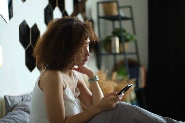 Sakin ol, pijamalı güzel Afrikalı kadın tek başına yatakta yatarken akıllı telefon kullanıyor..