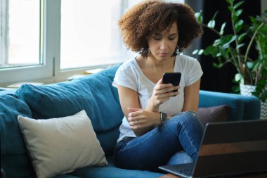 Genç Afro-Amerikalı iş kadını elinde akıllı telefon tutarken dizüstü bilgisayar kullanıyor.