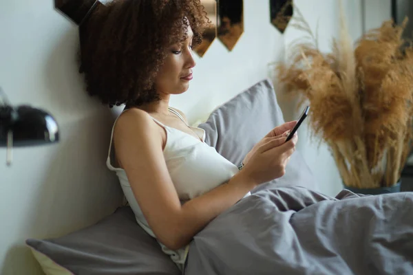 Black woman smiling and using smartphone on bed