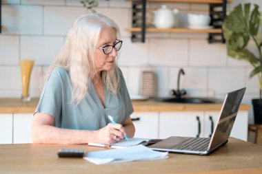 Üst düzey kadın mutfağında web sörfü yapmak için dizüstü bilgisayar kullanıyor. Kıdemli iş ve sosyal güvenlik kavramı. Olgun bir kadın iş yerinde oturmuş, bir ofiste defter bilgisayarı yazıyor..