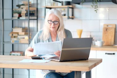 Üst düzey kadın mutfağında web sörfü yapmak için dizüstü bilgisayar kullanıyor. Kıdemli iş ve sosyal güvenlik kavramı. Olgun bir kadın iş yerinde oturmuş, bir ofiste defter bilgisayarı yazıyor..