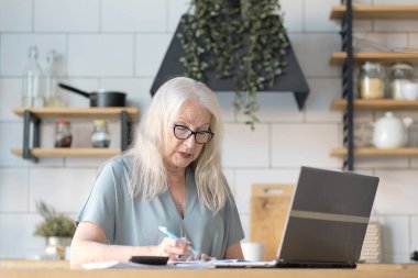 Üst düzey kadın mutfağında web sörfü yapmak için dizüstü bilgisayar kullanıyor. Kıdemli iş ve sosyal güvenlik kavramı. Olgun bir kadın iş yerinde oturmuş, bir ofiste defter bilgisayarı yazıyor..