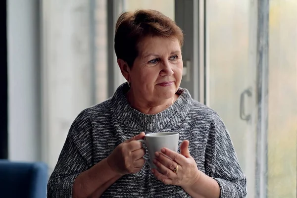 Portrait de femme âgée avec une tasse de café regardant par la fenêtre. Élégante dame mature buvant du café chaud et se relaxant près de la fenêtre souriant et rêvant à l'intérieur. — Photo