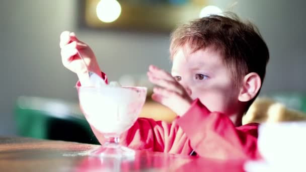 Um rapazinho a comer gelado no café. Criança bonita, desfrutando de iogurte de baunilha em um restaurante. tiro em câmera lenta. — Vídeo de Stock