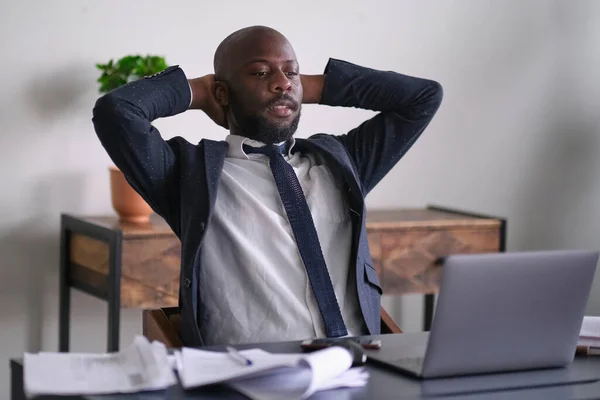 Relaxed worker finished work completed task taking break after hard workday putting head on hand. Thoughtful African American businessman looking out window sitting at office desk.