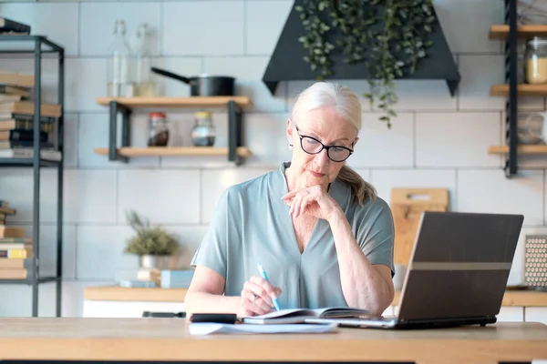 Mujer mayor que utiliza el ordenador portátil para navegar por Internet en su cocina. El concepto de empleo de alto nivel, seguridad social. Señora madura sentada en el trabajo escribiendo una computadora portátil en una oficina en casa. — Foto de Stock