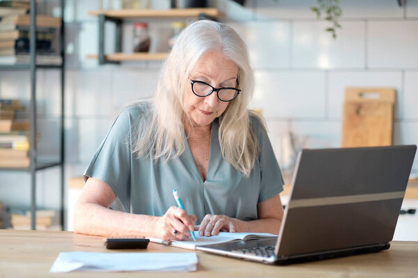Senior woman using laptop for websurfing in her kitchen. The concept of senior employment, social security. Mature lady sitting at work typing a notebook computer in an home office.