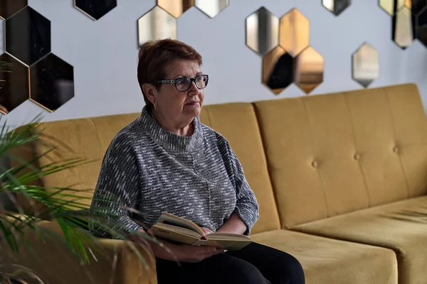 Hermosa anciana en ropa casual leyendo en el sofá en casa. Mujer mayor con mala vista en gafas mira en un libro. —  Fotos de Stock