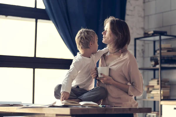 Young mom and son look at each other. Happy family in the morning. Beautiful woman with her son having breakfast in kitchen together, son looking at mother and smiling — Stock Photo, Image