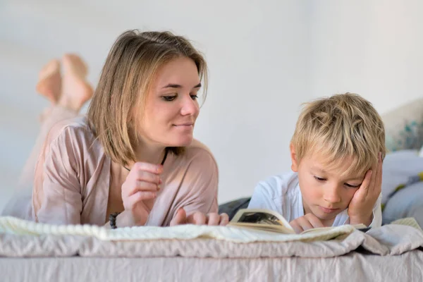 Máma učí syna číst knihu, je doma na posteli. Vzdělávací koncepty. Pastelové barvy, selektivní zaměření. — Stock fotografie