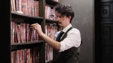 Eski karanlık bir kütüphanede klasik kitapları karıştıran şık giyimli bir adam. Manzarayı kapat. Yavaş çekim videosu. depolama görüntüleri