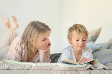 Anne oğluna evde yatağın üstünde kitap okumayı öğretiyor. Eğitim Konseptleri. Pastel renkler, seçici odak.