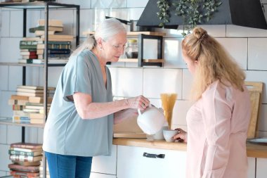 Arkadaş canlısı aile fotoğraflarına evde bakıyor. Kıdemli kadın kızıyla arayı kapatmanın tadını çıkarıyor. Onlar mutfakta bir fincan çay içiyorlar. Oturma odasında kadın bakıcısı olan yaşlı bir kadın.