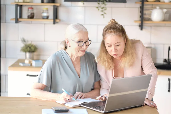 daughter helps her elderly mother figure it out online with her personal account. Woman teaching senior mother to use internet at home. Senior woman with her daughter looking at modern gadget indoors.