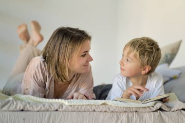 Anne oğluna evde yatağın üstünde kitap okumayı öğretiyor. Eğitim Konseptleri. Pastel renkler, seçici odak.