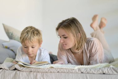 Anne oğluna evde yatağın üstünde kitap okumayı öğretiyor. Eğitim Konseptleri. Pastel renkler, seçici odak.
