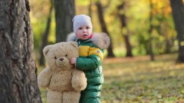 Küçük çocuk parkta pelüş bir oyuncakla oynuyor. Ilık ceketli komik bebek su samuru ve oyuncak ayısı. Manzarayı kapat. Yavaş çekim videosu. depolama görüntüleri