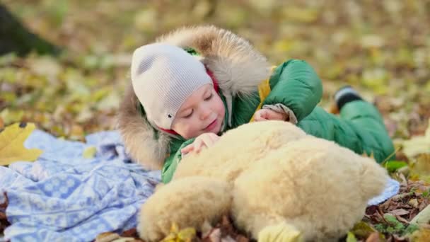 Menino brincando com um brinquedo de pelúcia no parque. engraçado bebê otty em um casaco quente e seu ursinho de pelúcia. vista de perto. Vídeo em câmara lenta. imagens de stock — Vídeo de Stock