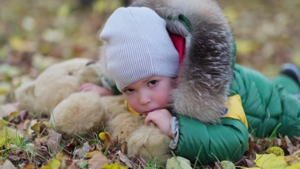 Маленький мальчик играет с плюшевой игрушкой в парке. Забавный малыш Отти в теплой куртке и его плюшевый мишка. close up view. Медленное видео. Отснятый материал — стоковое видео