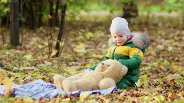 Un ragazzino che gioca con un peluche nel parco. divertente bambino otty in un caldo piumino e il suo orsacchiotto. vista da vicino. Video al rallentatore. stock di filmati — Video Stock