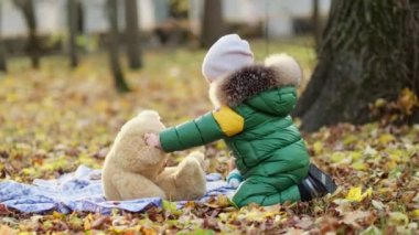 Küçük çocuk parkta pelüş bir oyuncakla oynuyor. Ilık ceketli komik bebek su samuru ve oyuncak ayısı. Manzarayı kapat. Yavaş çekim videosu. depolama görüntüleri
