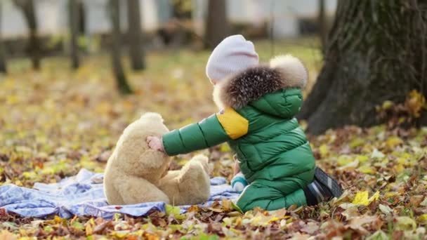 Menino brincando com um brinquedo de pelúcia no parque. engraçado bebê otty em um casaco quente e seu ursinho de pelúcia. vista de perto. Vídeo em câmara lenta. imagens de stock — Vídeo de Stock