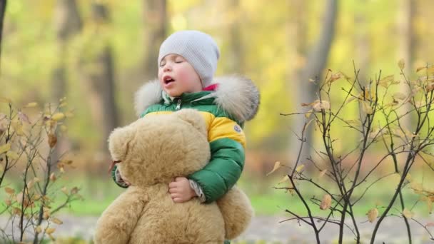 Un ragazzino che gioca con un peluche nel parco. divertente bambino otty in un caldo piumino e il suo orsacchiotto. vista da vicino. Video al rallentatore. stock di filmati — Video Stock