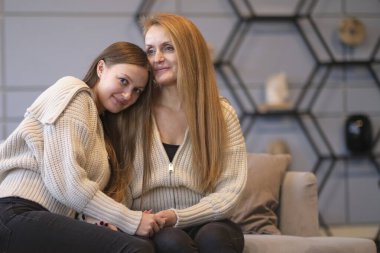 Neşeli genç bir kadın, yaşlı bir anneye sarılıyor. Oturma odasında dinlenirken koltukta oturuyor. Mutlu anne ve arkadaş canlısı kız el ele tutuşup birbirlerine bakıyorlar..