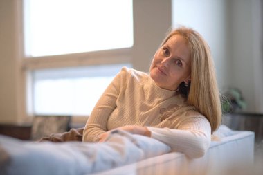 Evde gülümseyen mutlu kadın. Gülümseyen orta yaşlı sarışın kadın, ışıldayan bir gülümseme ile evdeki koltukta oturmuş kameraya bakıyor.