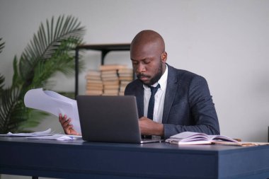 Genç Afrikalı yönetici ofisinde masasında otururken doküman okuyor ve dizüstü bilgisayarla çalışıyor. yumuşak odak.