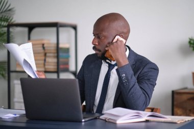 Genç Afrikalı yönetici ofisinde masasında otururken doküman okuyor ve dizüstü bilgisayarla çalışıyor. Çalışan ya da girişimci telefonla pazarlık yapıyor. yumuşak odak