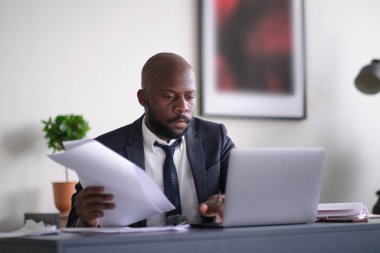 Genç Afrikalı yönetici ofisinde masasında otururken doküman okuyor ve dizüstü bilgisayarla çalışıyor. yumuşak odak.
