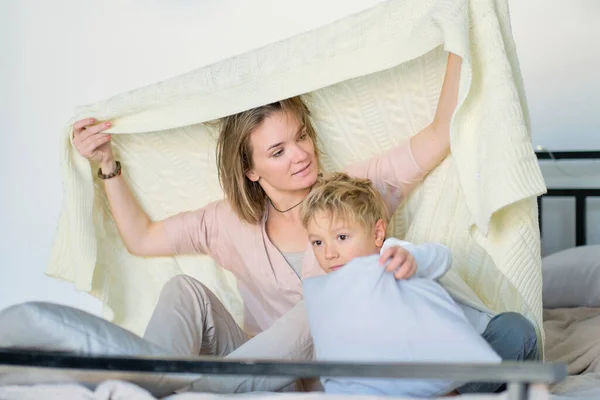 Máma a dítě se baví v ložnici na posteli, hrají si pod dekou. Mladá matka se svým synem oblečený hrát v posteli o víkendu spolu, líné ráno, teplé a útulné scény. — Stock fotografie
