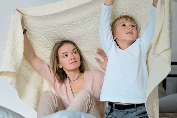 Máma a dítě se baví v ložnici na posteli, hrají si pod dekou. Mladá matka se svým synem oblečený hrát v posteli o víkendu spolu, líné ráno, teplé a útulné scény. — Stock fotografie