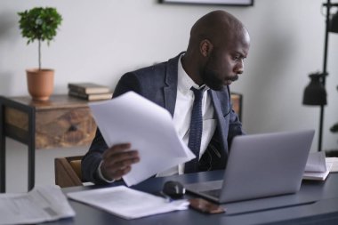 Genç Afrikalı yönetici ofisinde masasında otururken doküman okuyor ve dizüstü bilgisayarla çalışıyor. yumuşak odak.