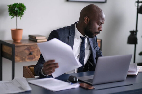 Young African executive reading documents and working on a laptop while sitting at his desk in an office. soft focus.