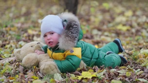 Un ragazzino che gioca con un peluche nel parco. divertente bambino otty in un caldo piumino e il suo orsacchiotto. vista da vicino. Video al rallentatore. stock di filmati — Video Stock