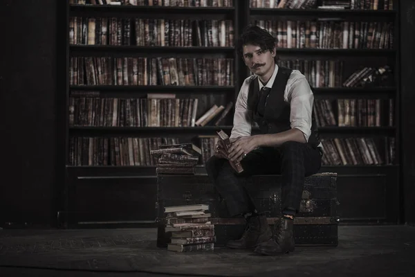 pensive man stands at night in a dark library room and holding a vintage book. stylish man with a mustache straightens the bracelet.