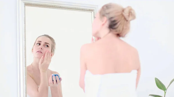 예쁜 여자가 아늑 한 욕실에서 거울 반사를 보면서 얼굴에 크림을 바르는 모습. — 스톡 사진