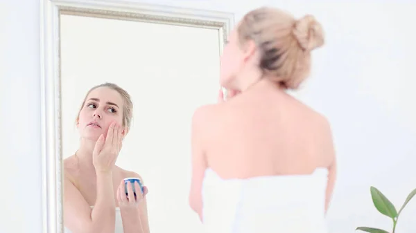 Pretty female applying cream on face while looking at mirror reflection in cozy bathroom.