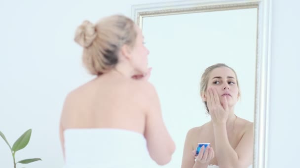 Dame appliquant crème hydratante lotion sur les joues, la finition matin routine de soins de la peau domestique. Femme souriante se toilettant après la douche dans la salle de bain. — Video