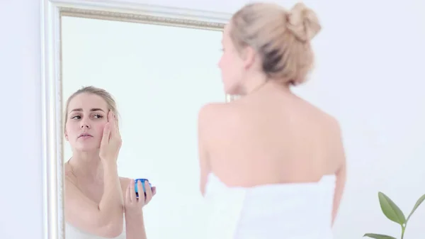Pretty female applying cream on face while looking at mirror reflection in cozy bathroom.