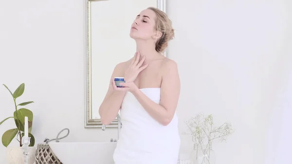 Dame appliquant crème hydratante lotion sur les joues, la finition matin routine de soins de la peau domestique. Femme souriante se toilettant après la douche dans la salle de bain. — Photo