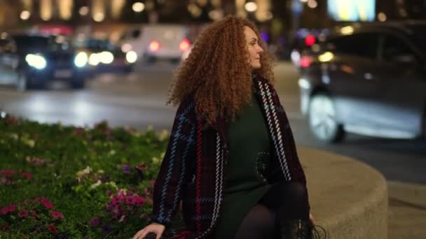 Retrato de una hermosa joven descansando en un banco en el centro de la ciudad, en el fondo los coches están pasando y las luces de la ciudad están brillando. uno en una gran ciudad. — Vídeos de Stock