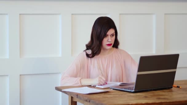 Linda joven sentada en el escritorio y escribiendo en el portátil y tareas de escritura para el día en un cuaderno. — Vídeos de Stock