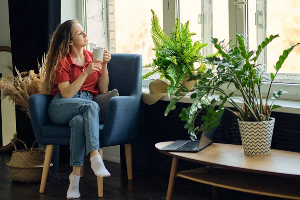 Fiatal nő egy csésze kávéval a kezében ül egy székben az ablak mellett egy hangulatos nappaliban — Stock Fotó