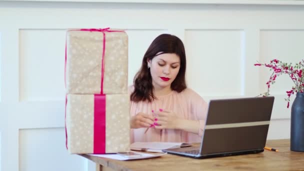 Jovencita bonita sentada en el escritorio y escribiendo en el portátil y hablando con iconos de cajas de regalo. caja recibe buena compra de la tienda en línea en casa, concepto de envío de correo postal — Vídeos de Stock