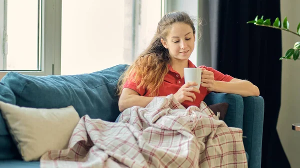Fiatal nő ül a kanapén, takaróval borítva, kezében egy csésze kávét tart. Lány otthon.. — Stock Fotó