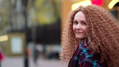 Güzel genç bir kadın kameraya bakıyor ve neşeyle gülümsüyor. Gün ışığında sokakta gülen surat. Dışarda olumlu bir portre.